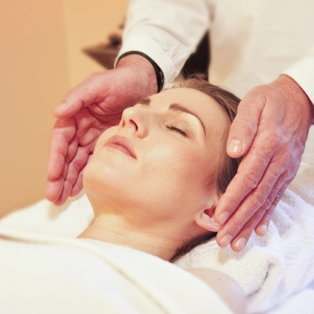 una mujer tumbada boca arriba y dos manos a los costados de su cara dando Reiki