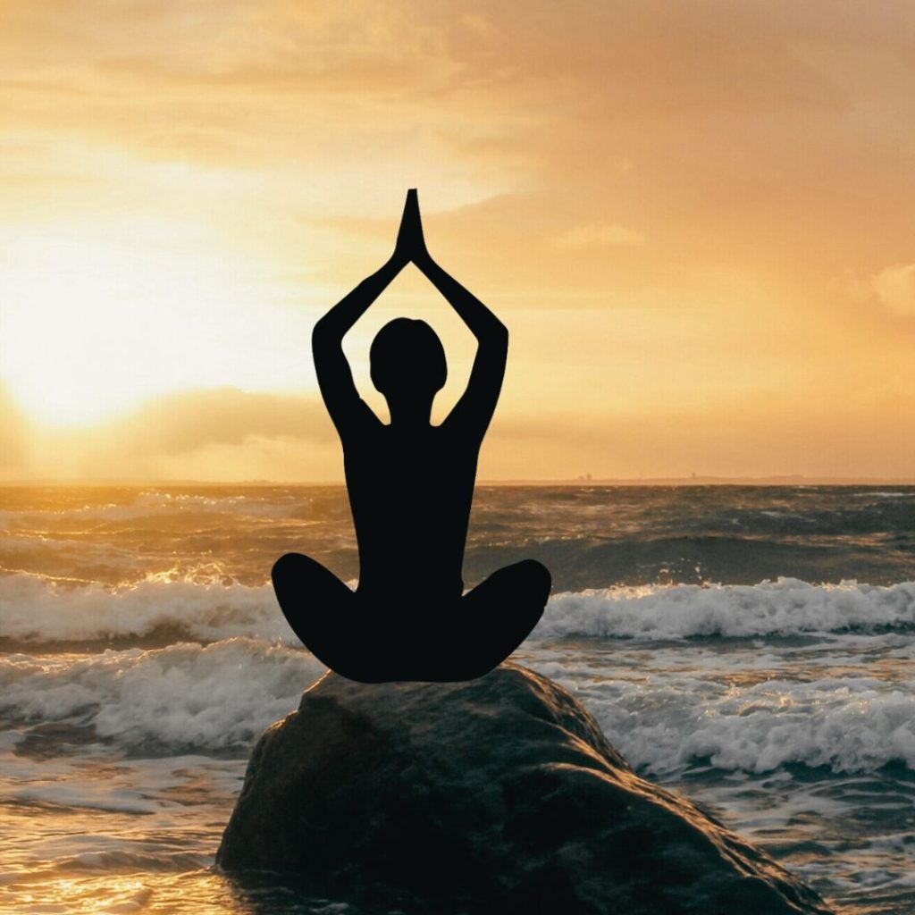 Una silueta de mujer en postura de yoga sentada sobre una roca dentro del mar en el anochecer