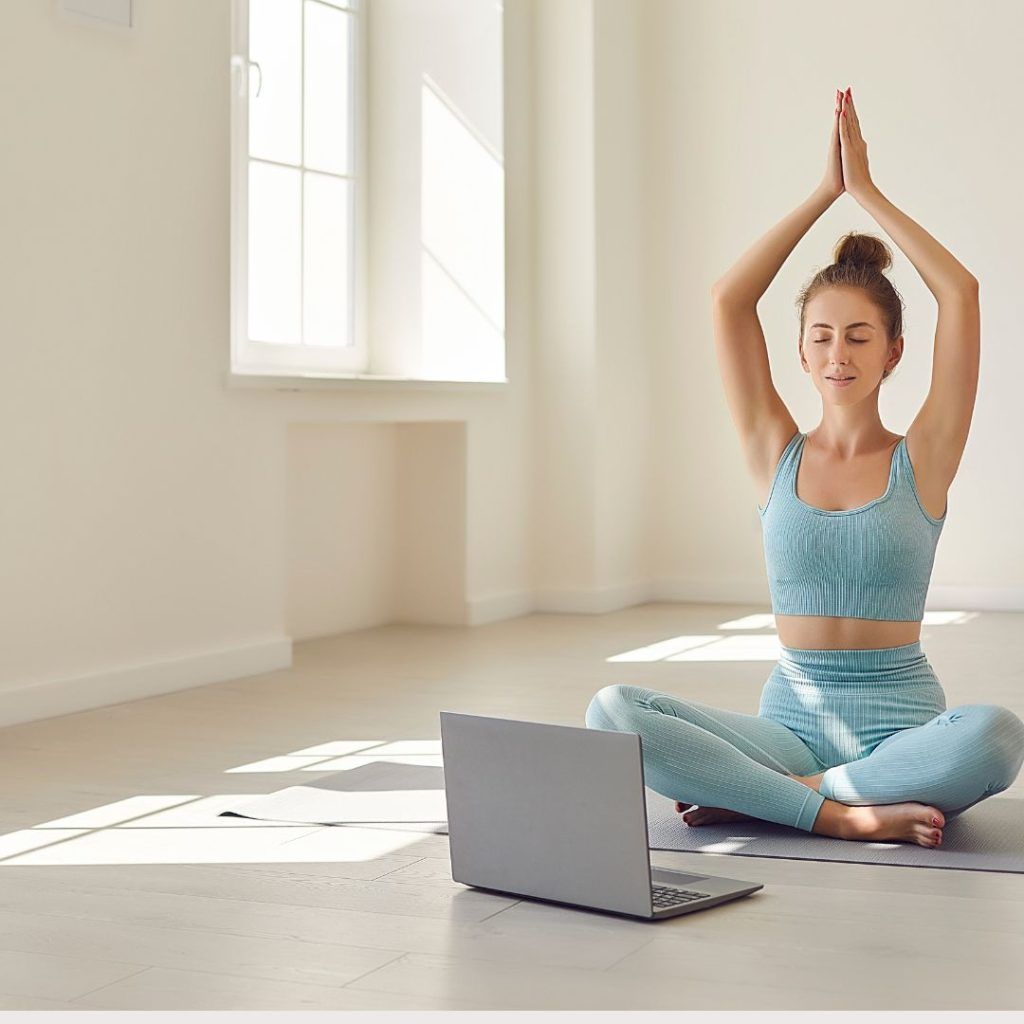 Una mujer en ropa de deporte, sentada con las piernas cruzadas y con los brazos levantados en postura de Yoga frente a un ordenador