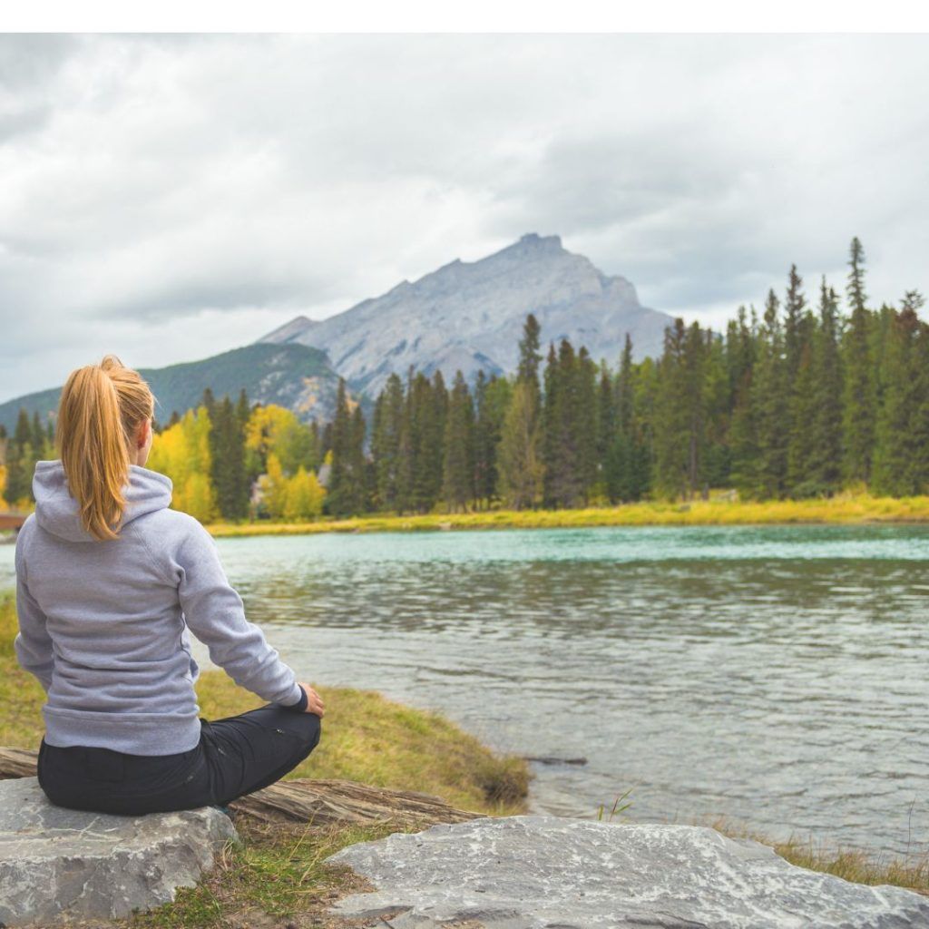 una mujer medita junto a un lago con una montaña y un bosque al fondo. Meditación en swadisthana chakra el agua de la vida
